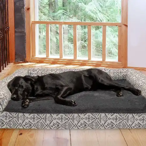 A large black dog lies stretched out on a gray orthopedic sofa-style dog bed with patterned side bolsters, placed on a wooden floor near a window, resting comfortably with full body support.